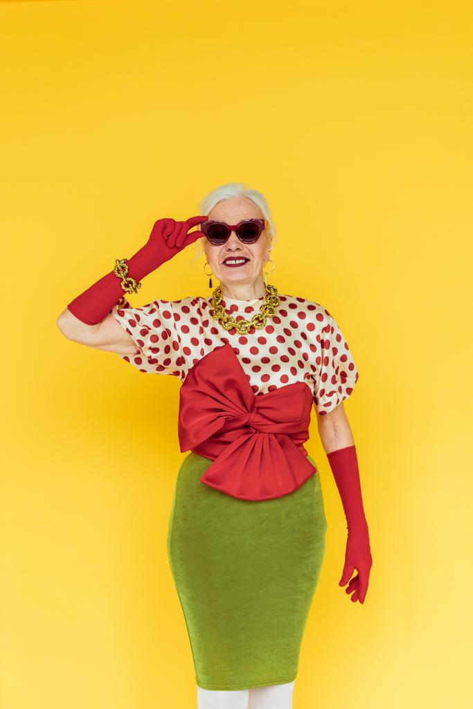 Elegante donna anziana sorridente e tenendo gli occhiali da sole isolati sul giallo
 - Foto, immagini