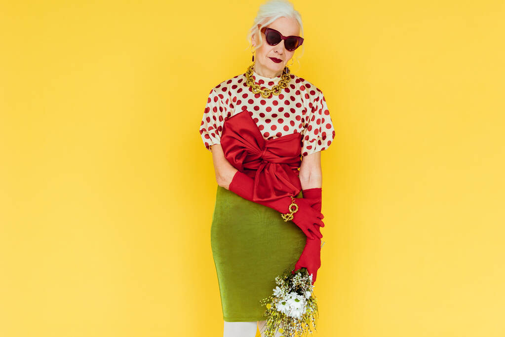 Beautiful elderly woman in sunglasses holding bouquet of wildflowers isolated on yellow - Photo, Image