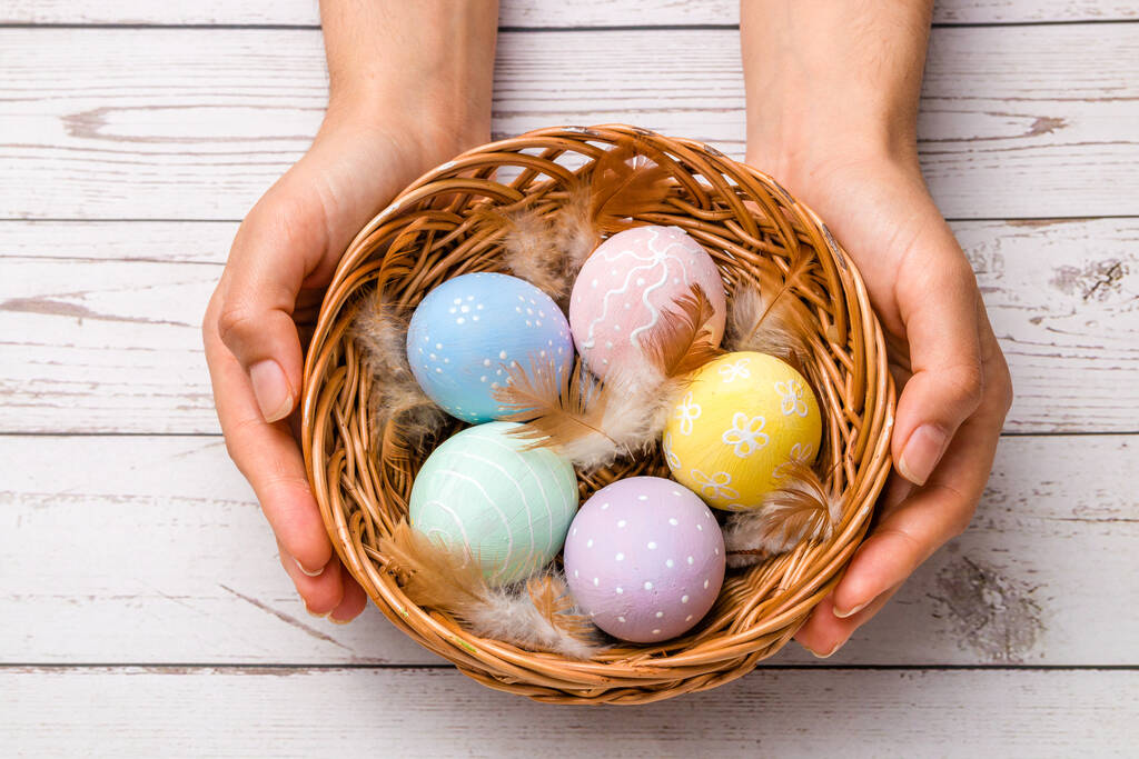 easter concept. closeup beautiful woman hands holding a wicker basket with hand-painted easter eggs in tender pastel colors and feather over wooden table. - Photo, Image