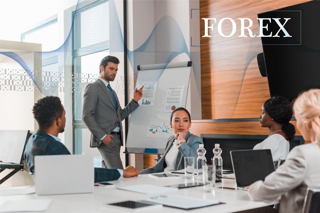 handsome businessman pointing with finger at flipchart with infographics while multicultural colleagues sitting at desk in conference hall, forex illustration - Photo, Image