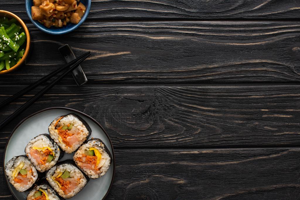 верхний вид палочек рядом с тарелкой с вкусной Джимбап и корейской гарнир на деревянной поверхности
  - Фото, изображение