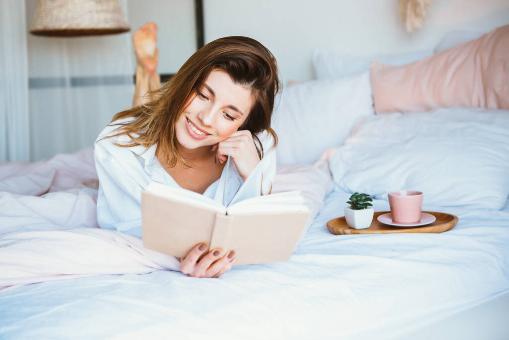 Ragazza che legge un libro, beve caffè o tè, riposa a letto
. - Foto, immagini