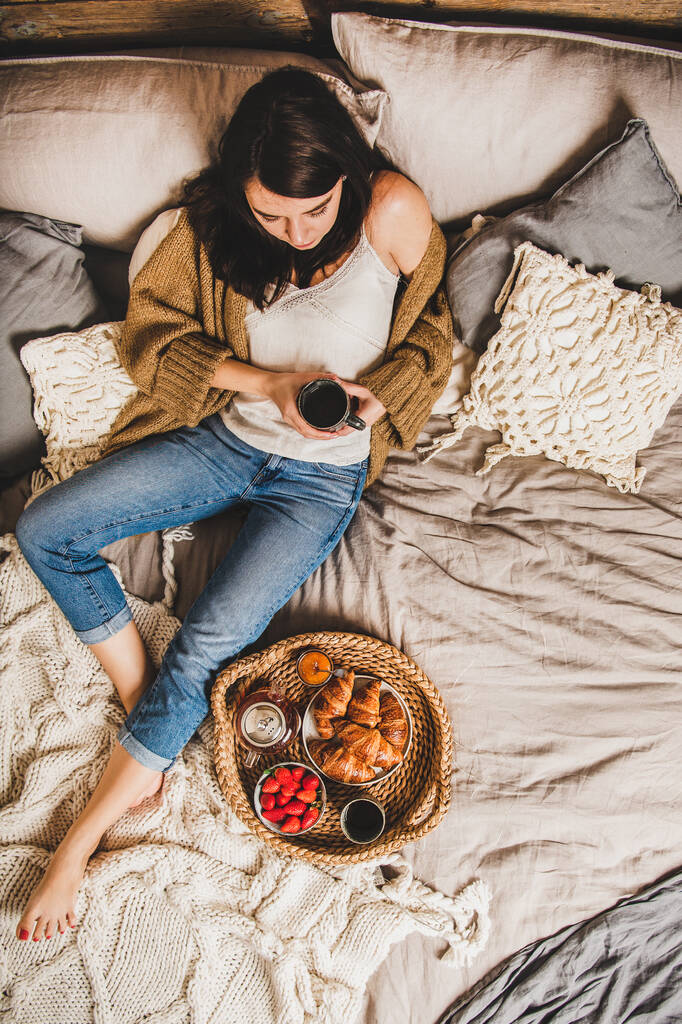 Mladá brunetka v neformálním oblečení sedí na pastelově zbarvených přikrývkách bosá a snídá v posteli s čerstvými croissanty, jahodami a čajem, výhled shora. Pojem pohodlného bydlení - Fotografie, Obrázek