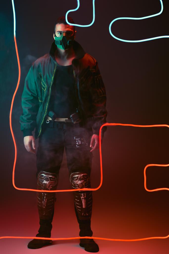 Mixed Race Cyberpunk-Spieler in Schutzmaske steht neben roter Neonbeleuchtung auf schwarz  - Foto, Bild
