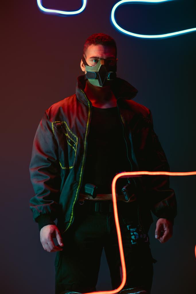 Bewaffneter und zweirassiger Cyberpunk-Spieler in Schutzmaske, der in der Nähe von Neonlicht auf Schwarz steht  - Foto, Bild
