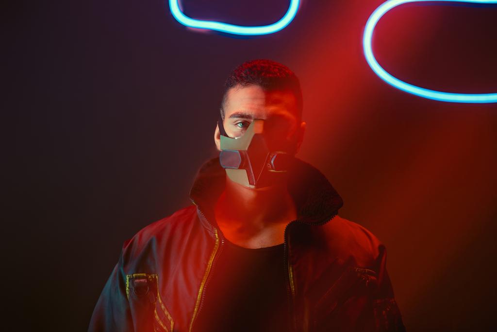 bi-raciale cyberpunk speler in beschermend masker kijkend naar camera in de buurt van neon verlichting op zwart  - Foto, afbeelding