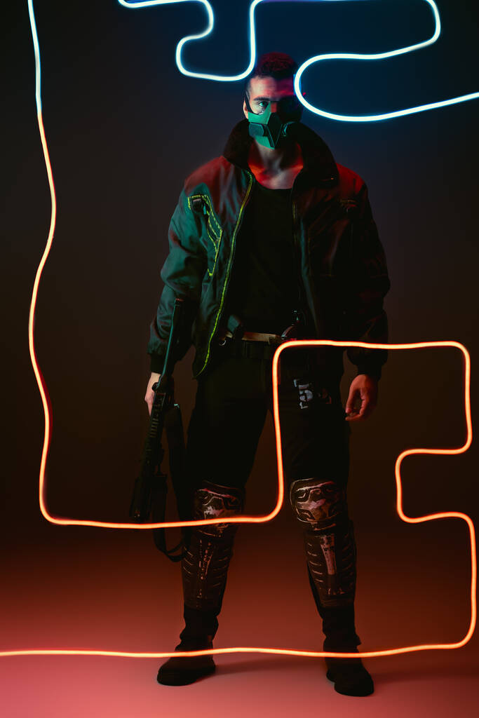 μικτή φυλή Cyberpunk player σε προστατευτική μάσκα κρατώντας όπλο, ενώ στέκεται κοντά νέον φωτισμό σε μαύρο  - Φωτογραφία, εικόνα