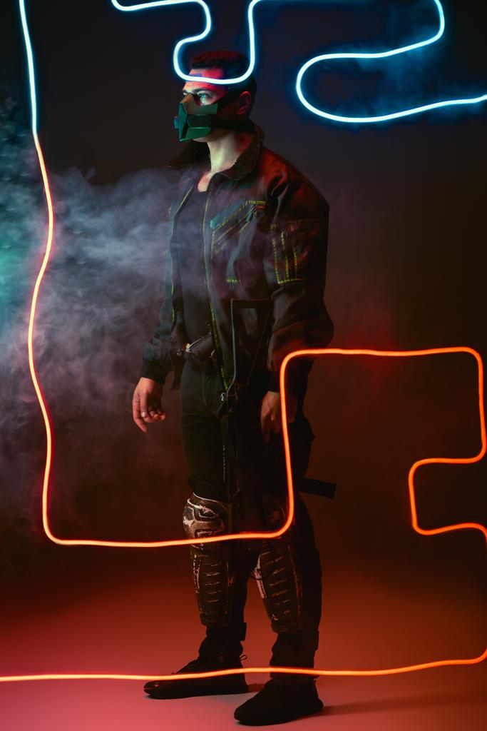 gewapende gemengde ras cyberpunk speler in beschermende masker staan in de buurt van neon verlichting op zwart met rook  - Foto, afbeelding