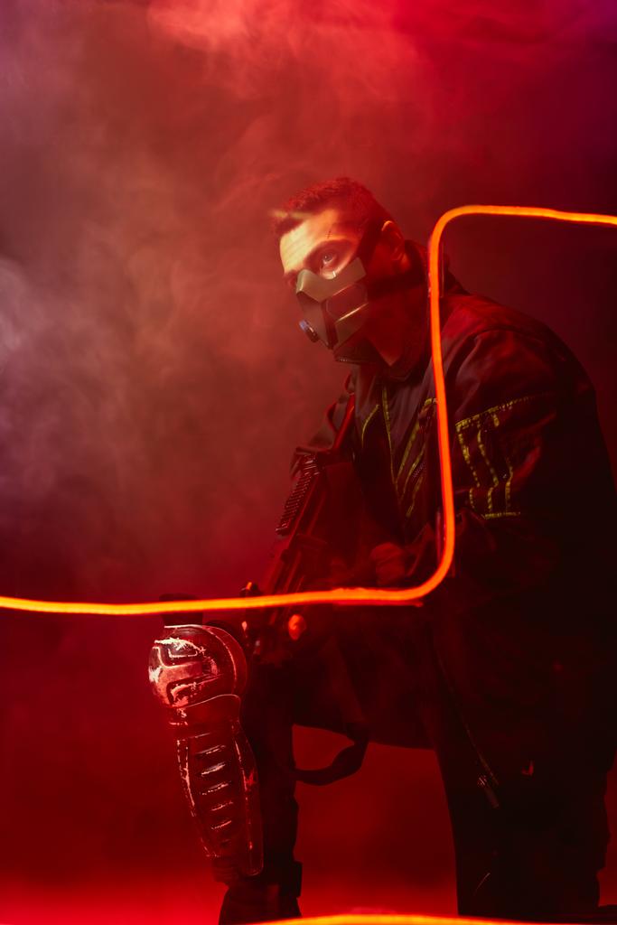 Koruyucu maskeli tehlikeli bir çift ırklı siber punk oyuncusunun elinde siyah ışıkta dumanlı bir silah var.  - Fotoğraf, Görsel