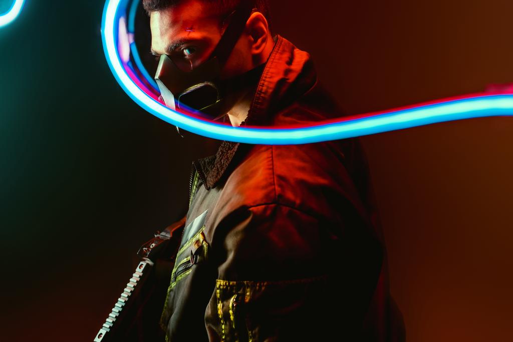 selektywne skupienie niebezpiecznej rasy mieszanej cyberpunk gracz w masce trzymając pistolet w pobliżu niebieskiego neonu oświetlenia na czarno  - Zdjęcie, obraz
