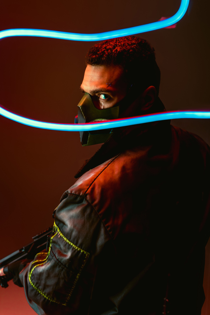 enfoque selectivo de jugador bi-racial cyberpunk en máscara protectora sosteniendo pistola cerca de la iluminación de neón azul en negro
  - Foto, imagen