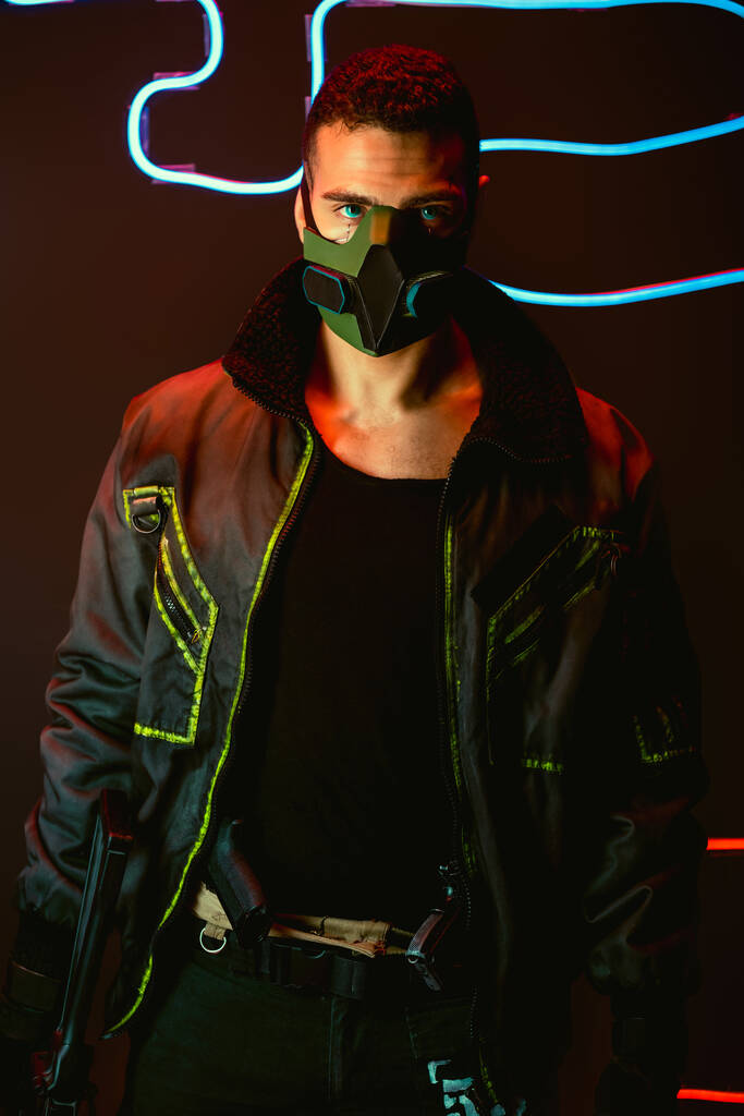 bi-raciale cyberpunk speler in beschermende masker holding gun in de buurt van blauwe neon verlichting op zwart  - Foto, afbeelding