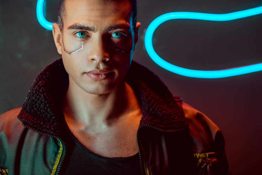 handsome and bi-racial cyberpunk man looking at camera near neon lighting on black  - Photo, Image