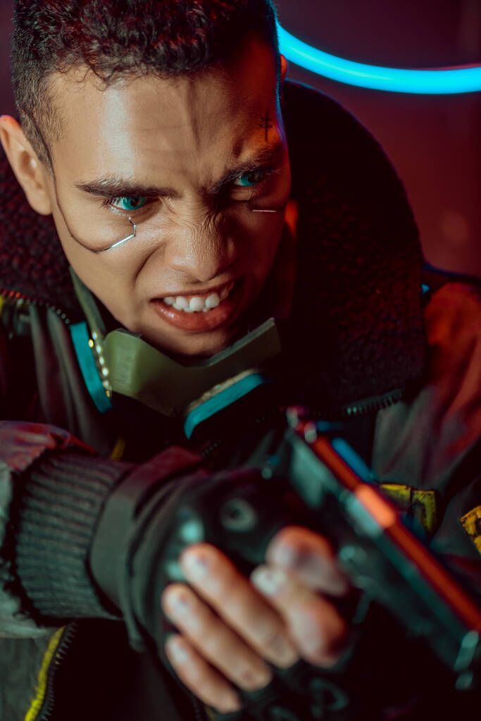 selectieve focus van boze bi-raciale cyberpunk speler met metalen platen op gezicht holding gun  - Foto, afbeelding