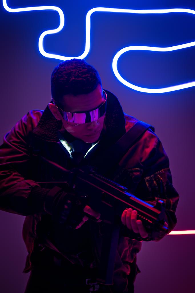uzbrojony mieszane wyścigu cyberpunk gracz w futurystyczne okulary trzymając pistolet w pobliżu neon oświetlenia  - Zdjęcie, obraz