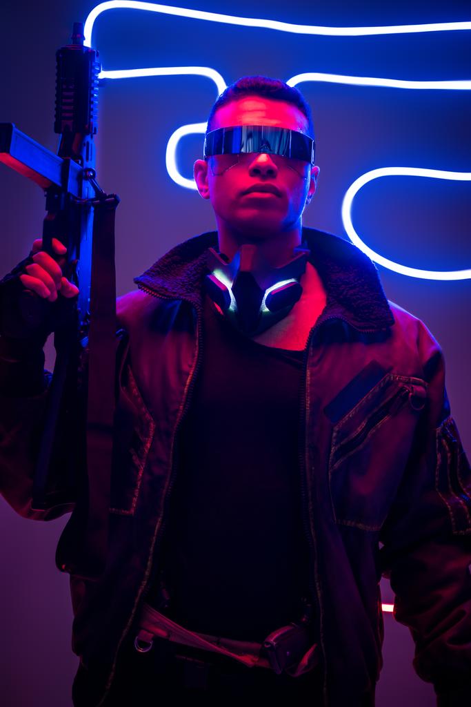 peligroso jugador cyberpunk de raza mixta en gafas futuristas con pistola cerca de la iluminación de neón
  - Foto, imagen