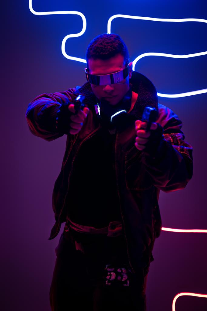 jóképű vegyes fajú cyberpunk játékos futurisztikus szemüvegben fegyvert tart neon világítás közelében  - Fotó, kép