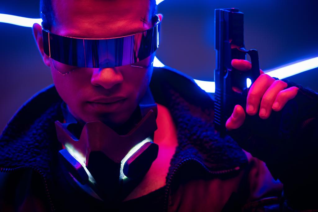 όμορφος παίκτης mixed race cyberpunk σε φουτουριστικό γυαλιά κρατώντας όπλο κοντά σε μπλε νέον φωτισμό  - Φωτογραφία, εικόνα