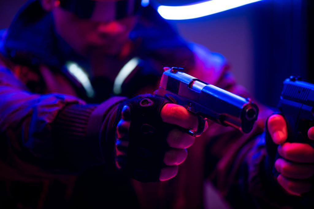επιλεκτική εστίαση των bi-φυλετικών cyberpunk player κρατώντας όπλα κοντά σε νέον φωτισμό  - Φωτογραφία, εικόνα