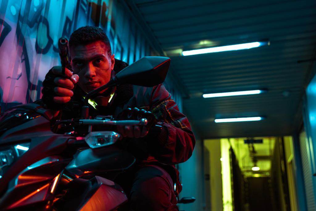 dangerous and mixed race cyberpunk player on motorcycle aiming gun on street with graffiti - Photo, Image