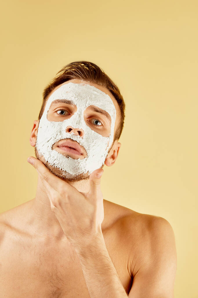 A fehér maszkos, jóképű, szakállas fiatalember portréja megérinti arcát a stúdióban, sárga alapon elszigetelve, férfi bőrápolási koncepcióval. - Fotó, kép