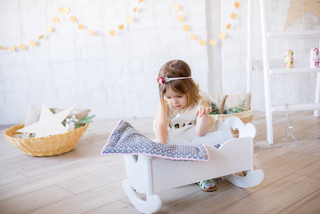 Pieni söpö tyttö valkoisessa mekossa pelaa nukkesängyssä kirkkaassa lastenhuoneessa, koristeltu leluilla ja seppeleillä.
. - Valokuva, kuva