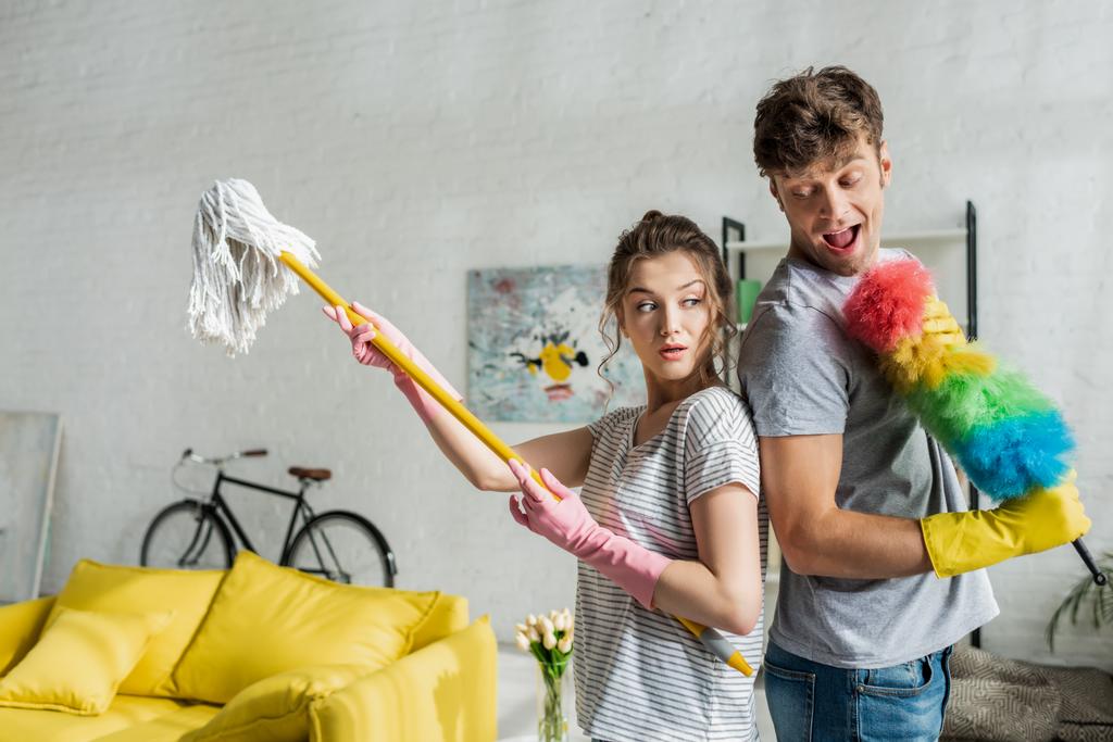 hombre excitado y hermosa mujer sosteniendo fregona y cepillo de plumero en casa
  - Foto, imagen