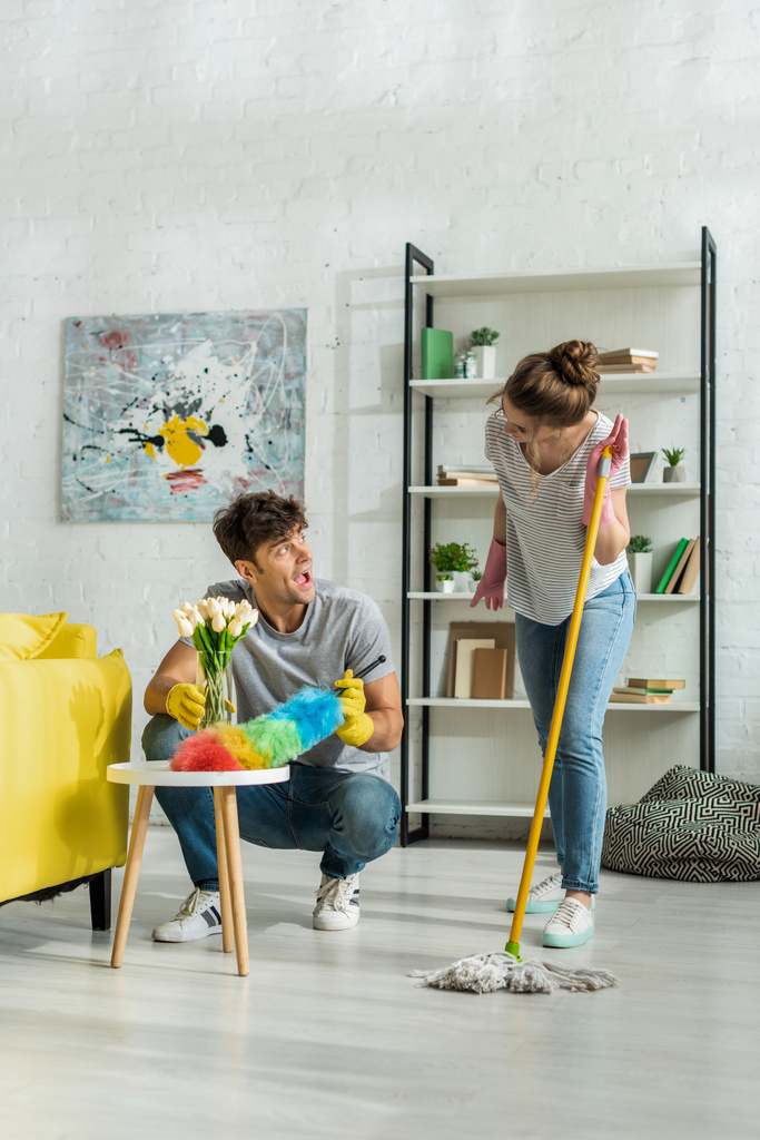 excited man and happy woman looking at each other while cleaning living room  - Photo, Image