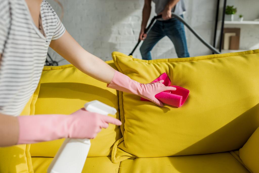 bijgesneden weergave van meisje wassen sofa met doek in de buurt van man schoonmaken woonkamer met stofzuiger - Foto, afbeelding