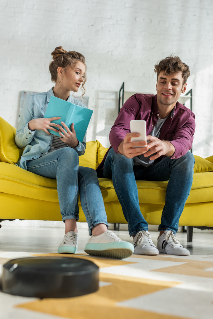 selektivní zaměření šťastný muž pomocí smartphone a žena drží knihu, zatímco robotický vysavač prací koberec v obývacím pokoji  - Fotografie, Obrázek