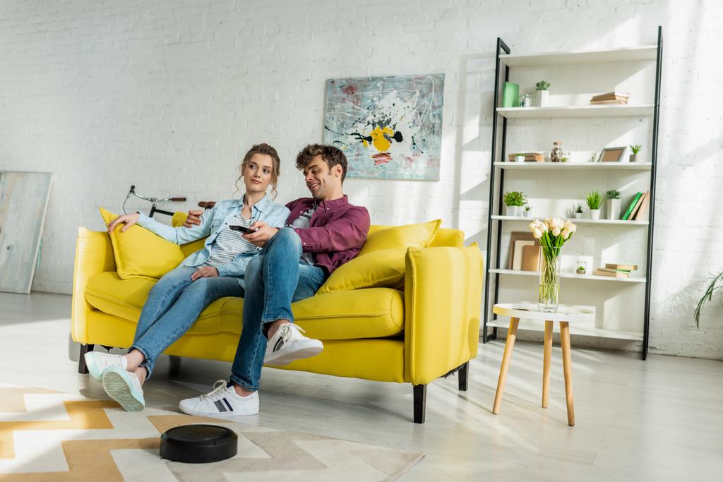 happy man and young woman watching movie while robotic vacuum cleaner washing carpet in living room  - Photo, Image
