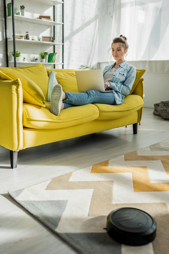 donna attraente utilizzando il computer portatile sul divano mentre robot aspirapolvere tappeto di lavaggio in soggiorno
  - Foto, immagini