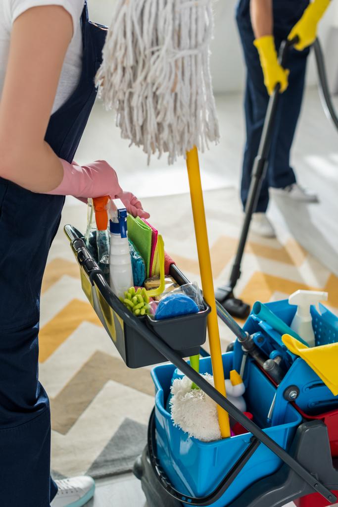 vista recortada de la limpiadora en guantes de goma de pie cerca del carro de limpieza y compañero de trabajo con aspiradora
  - Foto, imagen