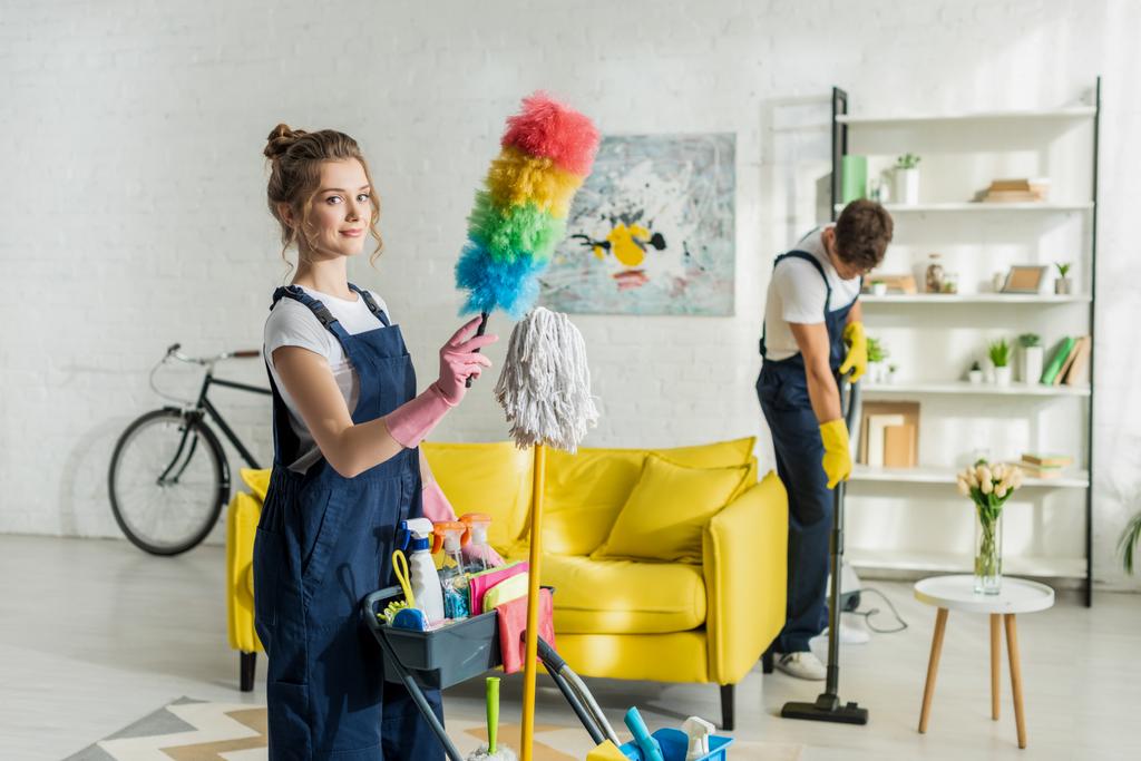 foyer sélectif des jeunes et heureux nettoyeur tenant brosse à poussière près du chariot de nettoyage
 - Photo, image