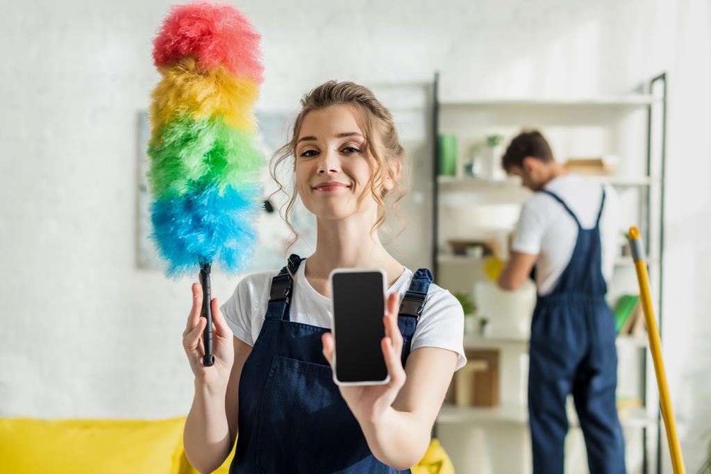 Boş ekran ve toz fırçası ile akıllı telefonu tutan mutlu ve çekici temizlikçinin seçici odak noktası - Fotoğraf, Görsel
