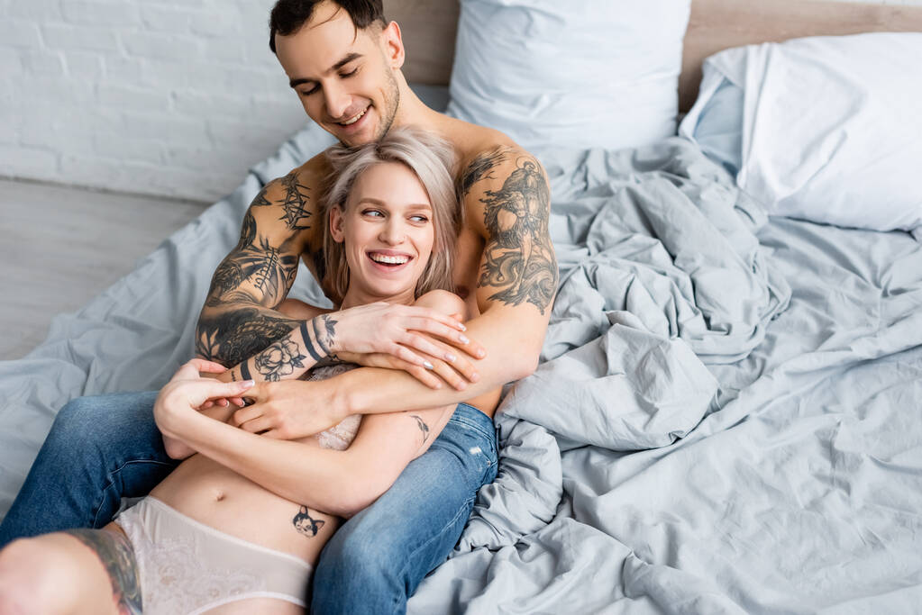 Hezký tetovaný muž objímající usmívající se přítelkyně ve spodním prádle na posteli  - Fotografie, Obrázek