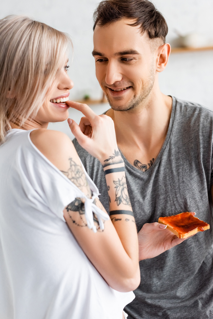 Knappe man glimlachen terwijl kijken naar getatoeëerde vriendin met toast in de keuken  - Foto, afbeelding