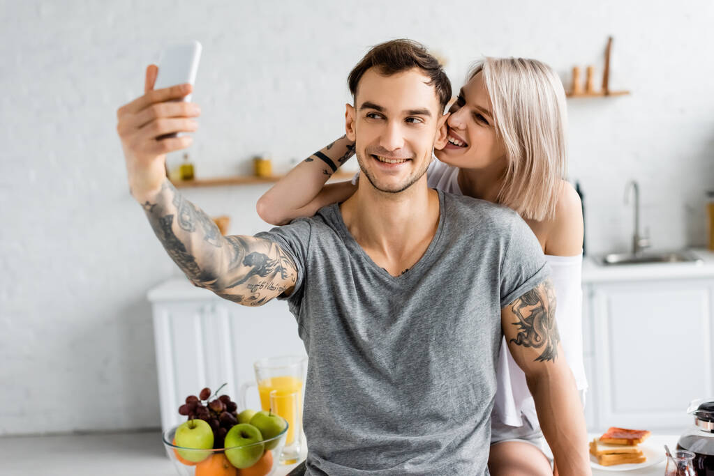 Focus sélectif de l'homme tatoué prenant selfie avec smartphone près de petite amie souriante sur la table de cuisine
  - Photo, image