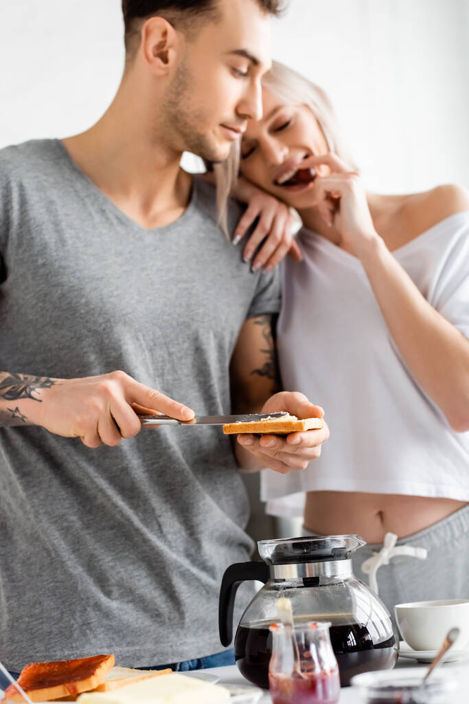 Szelektív fókusz tetovált férfi pirítóssal és késsel nézi mosolygó barátnője eszik szőlő mellett reggeli a konyhában  - Fotó, kép