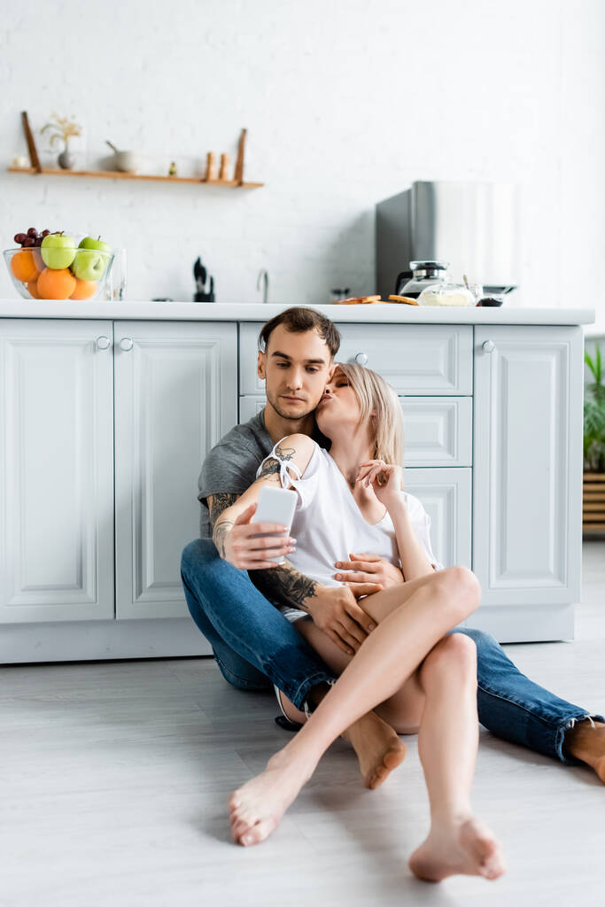 Menina bonita beijando namorado tatuado ao tomar selfie com smartphone no chão na cozinha
  - Foto, Imagem