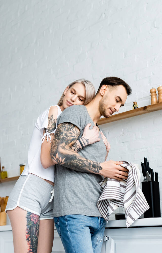Vista de ángulo bajo de la hermosa mujer abrazando novio tatuado con toalla en la cocina
  - Foto, imagen