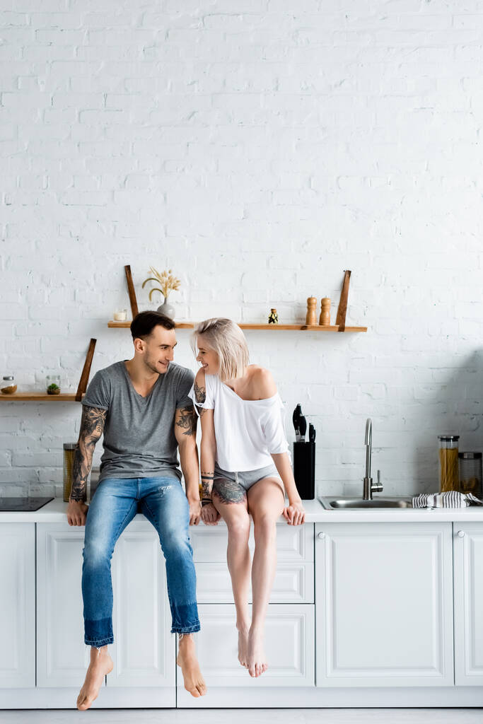 Tätowiertes Paar lächelt einander an, während es auf der Arbeitsplatte in der Küche sitzt  - Foto, Bild