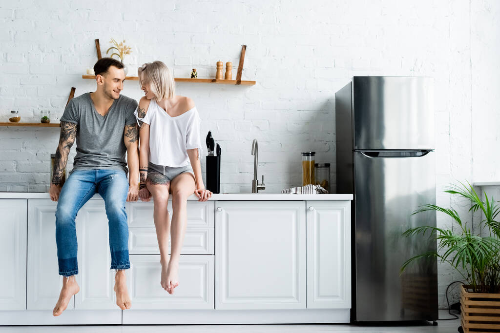 Tätowierter Mann lächelt schöne Freundin auf Küchenarbeitsplatte an  - Foto, Bild