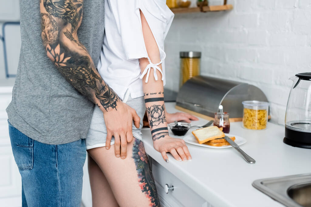 Καλλιεργημένη άποψη του άντρα με τατουάζ αγγίζοντας ισχίο της φίλης κατά τη διάρκεια του πρωινού στην κουζίνα  - Φωτογραφία, εικόνα