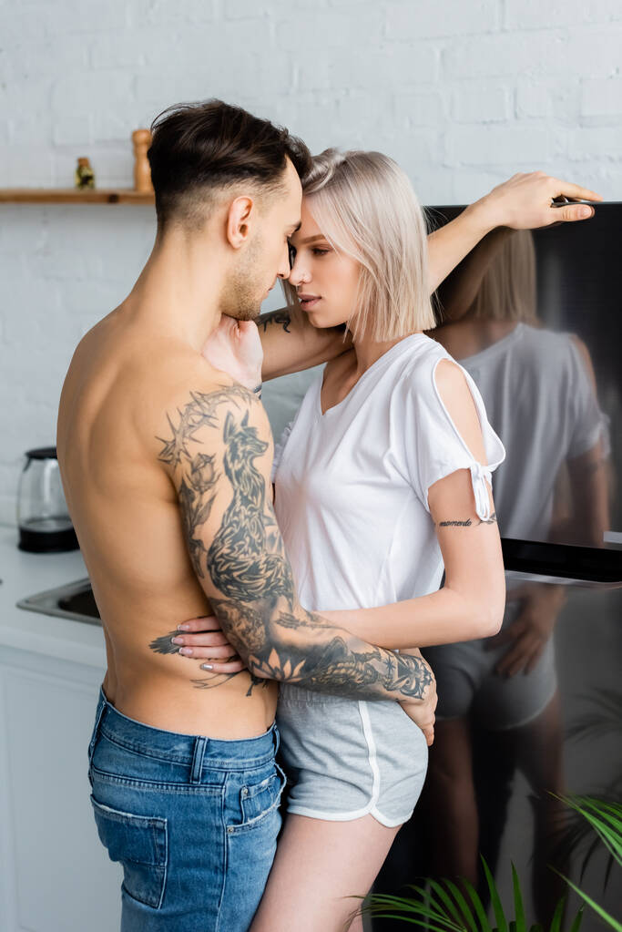Boční pohled na tetované dívky objímající bez košile přítele v blízkosti lednice v kuchyni  - Fotografie, Obrázek