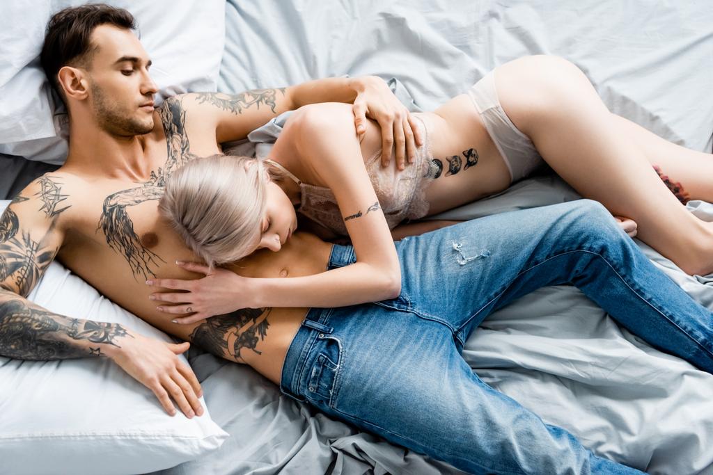 Bovenaanzicht van shirtloze getatoeëerde man omarmen mooi meisje in ondergoed op bed  - Foto, afbeelding