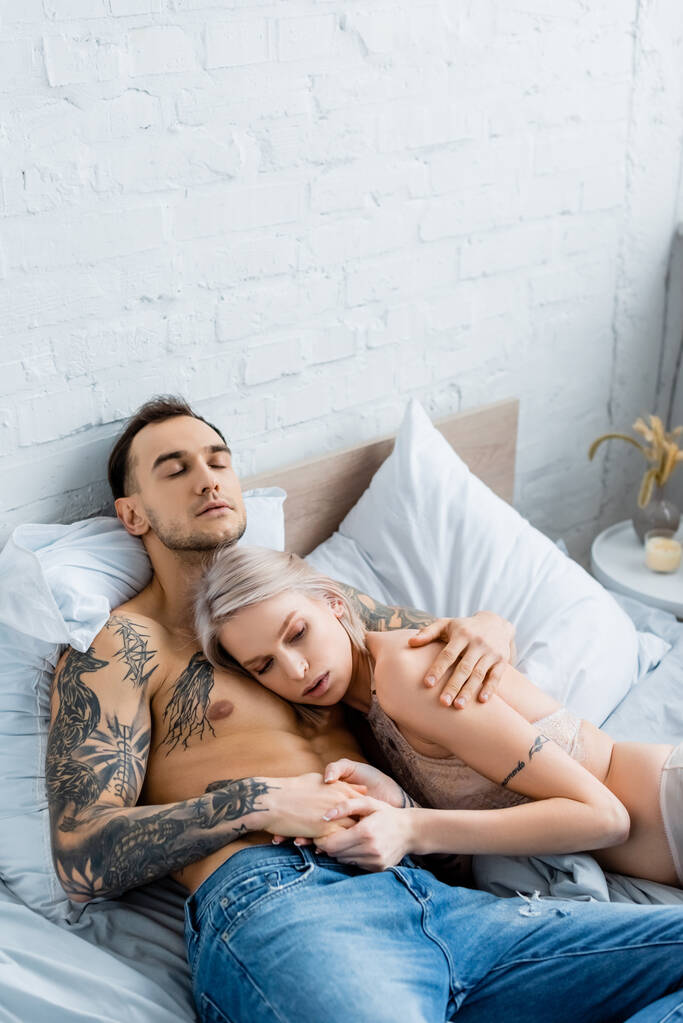 Atraktivní dívka ve spodním prádle dotýkající se ruky svalnatého tetovaného přítele na posteli  - Fotografie, Obrázek
