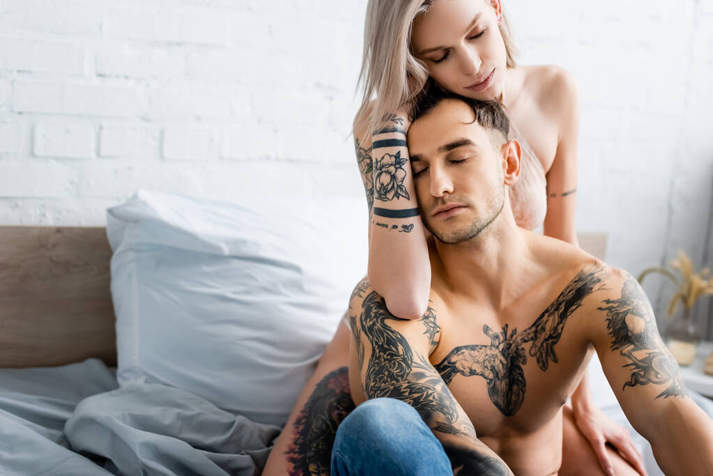Mulher sexy no sutiã abraçando sem camisa tatuado namorado na cama
  - Foto, Imagem