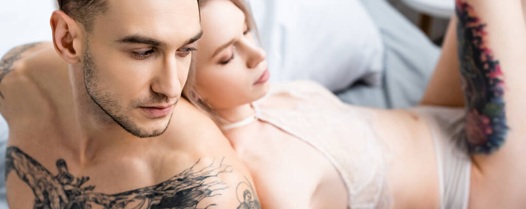 Panoramische shot van gespierde getatoeëerde man zitten op bed in de buurt van sexy vriendin in lingerie  - Foto, afbeelding