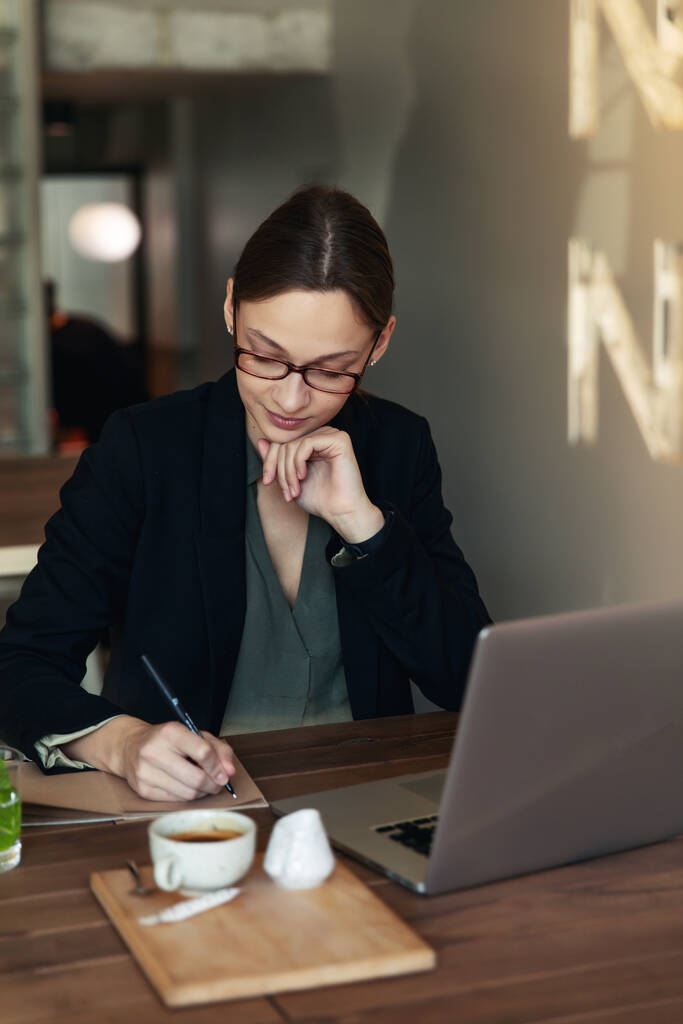 Siyah takım elbiseli ve gözlüklü bir iş kadını bir kafede oturuyor laptopun başında çalışıyor ve elinde plakları olan bir defter tutuyor. - Fotoğraf, Görsel